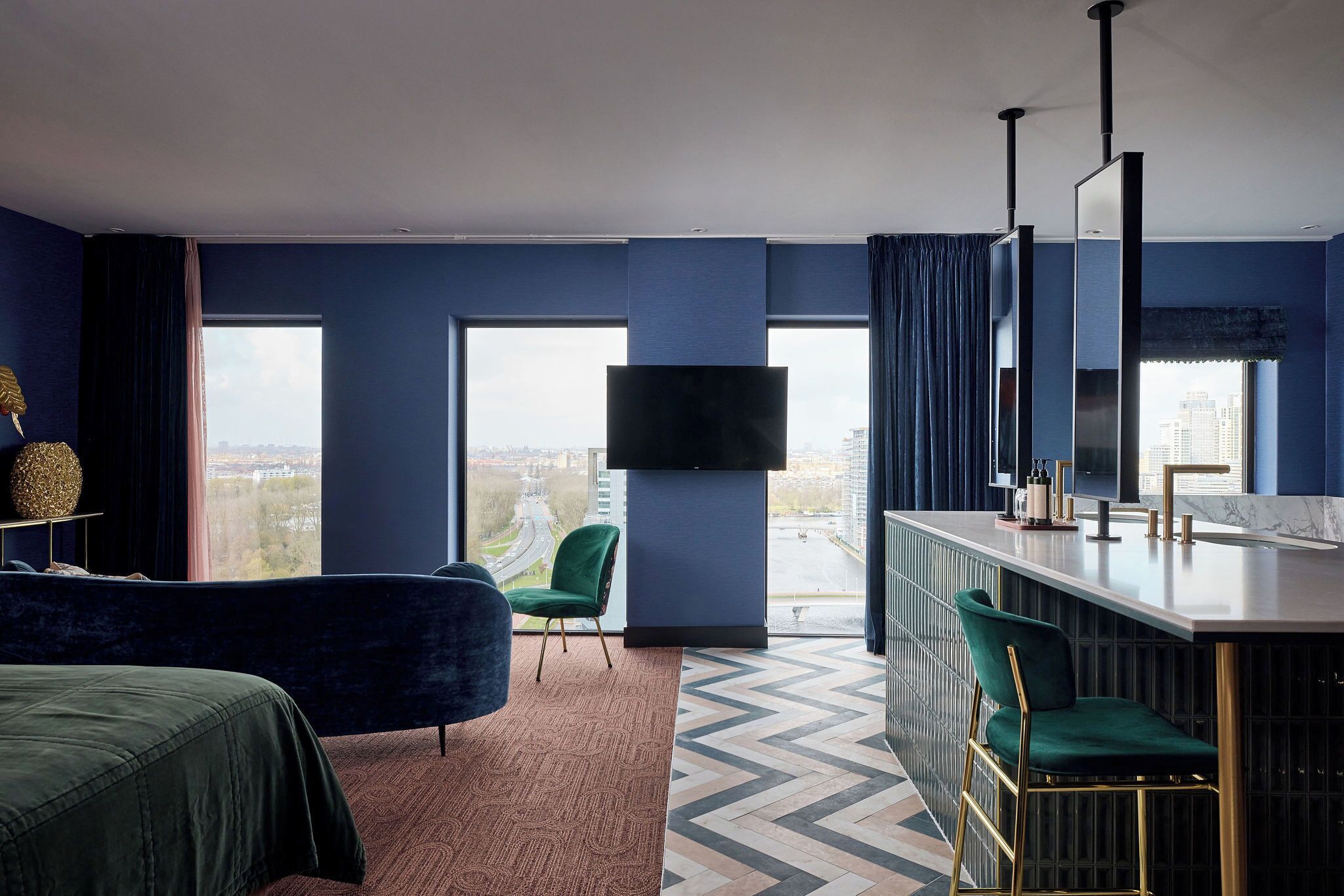 A modern hotel room with a large window, a flat-screen TV, a bar area, and a green chair.