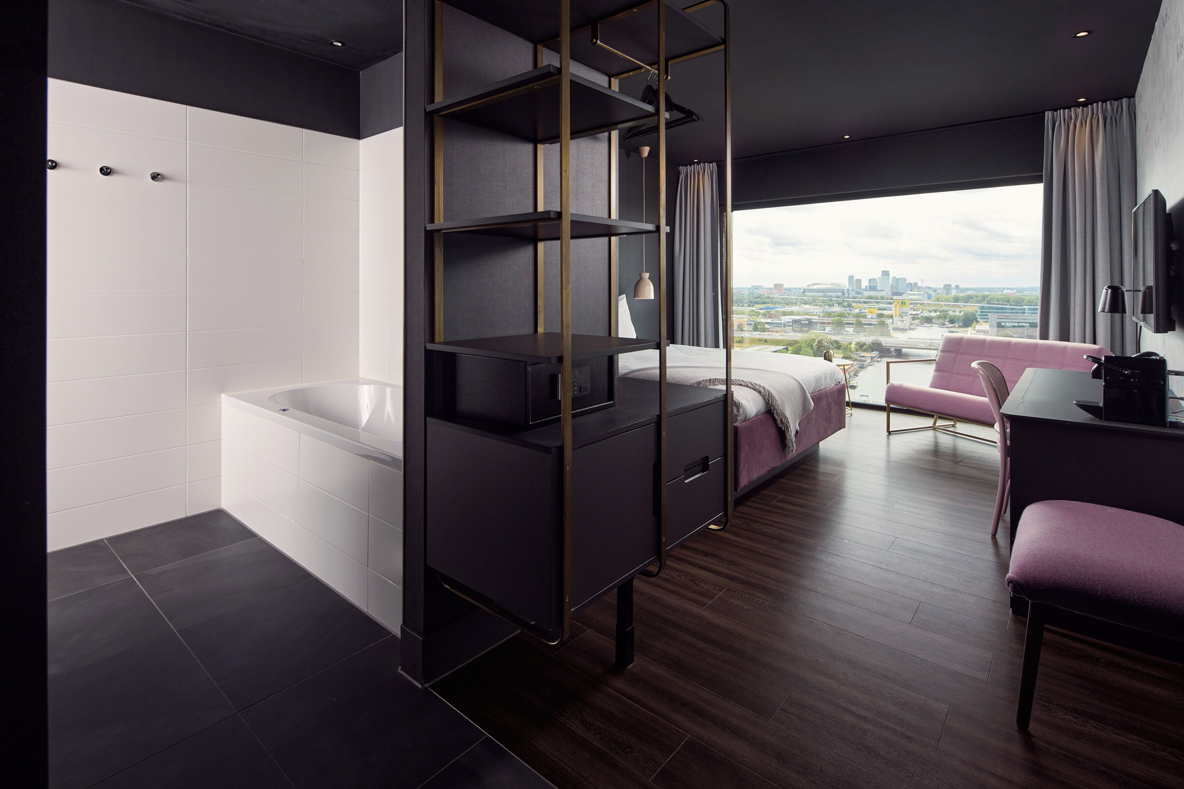Een moderne hotelkamer met een groot raam, een badkuip, een bureau en een roze stoel.