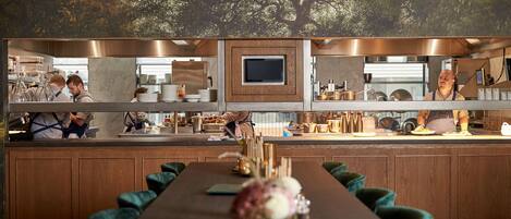 A restaurant with a long dining table, green upholstered chairs, and a mural of trees on the wall.