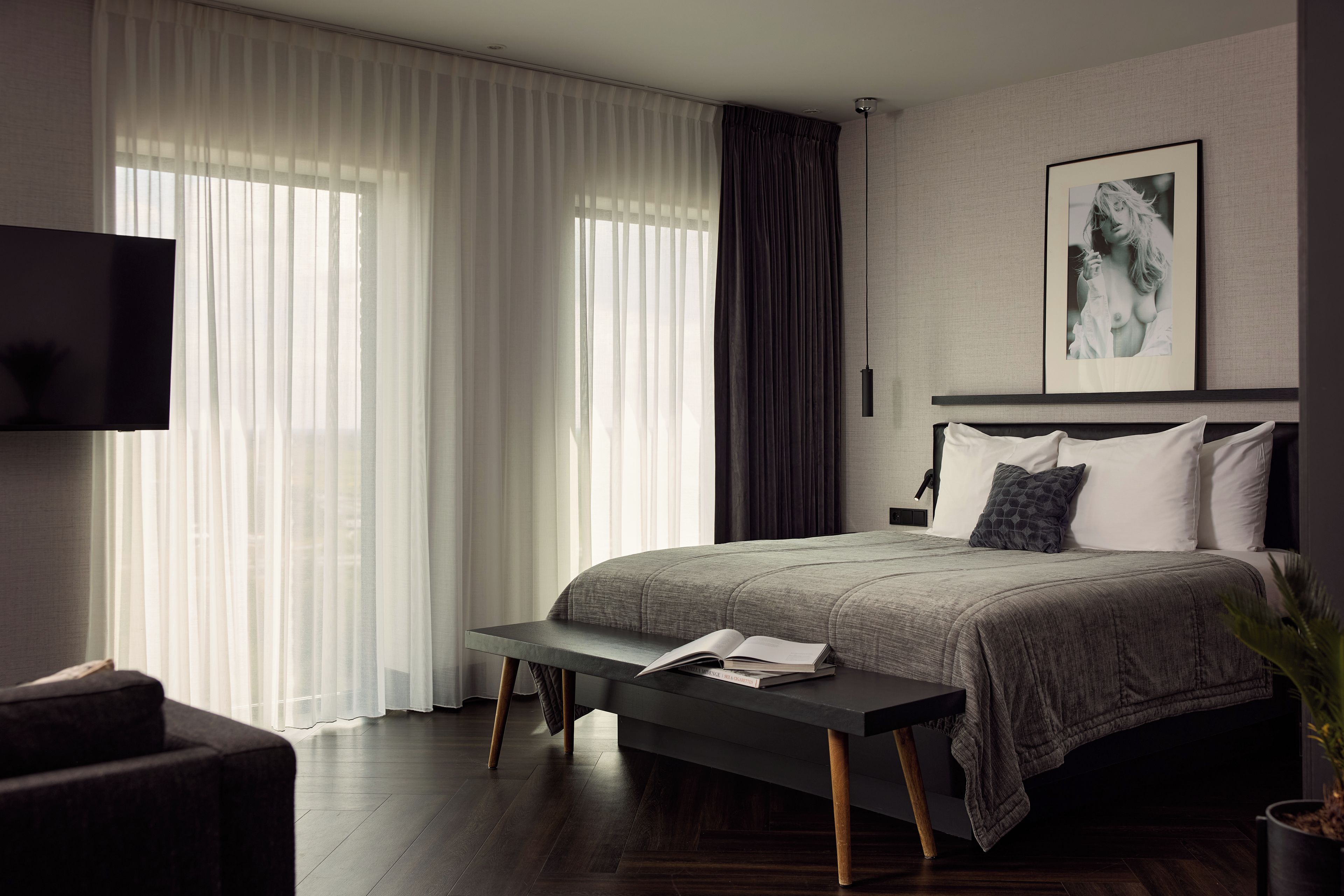 A modern bedroom with a large bed, a bedside table with a book, a TV mounted on the wall, and a window with sheer curtains.