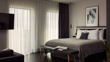 A modern bedroom with a large bed, a bedside table with a book, a TV mounted on the wall, and a window with sheer curtains.