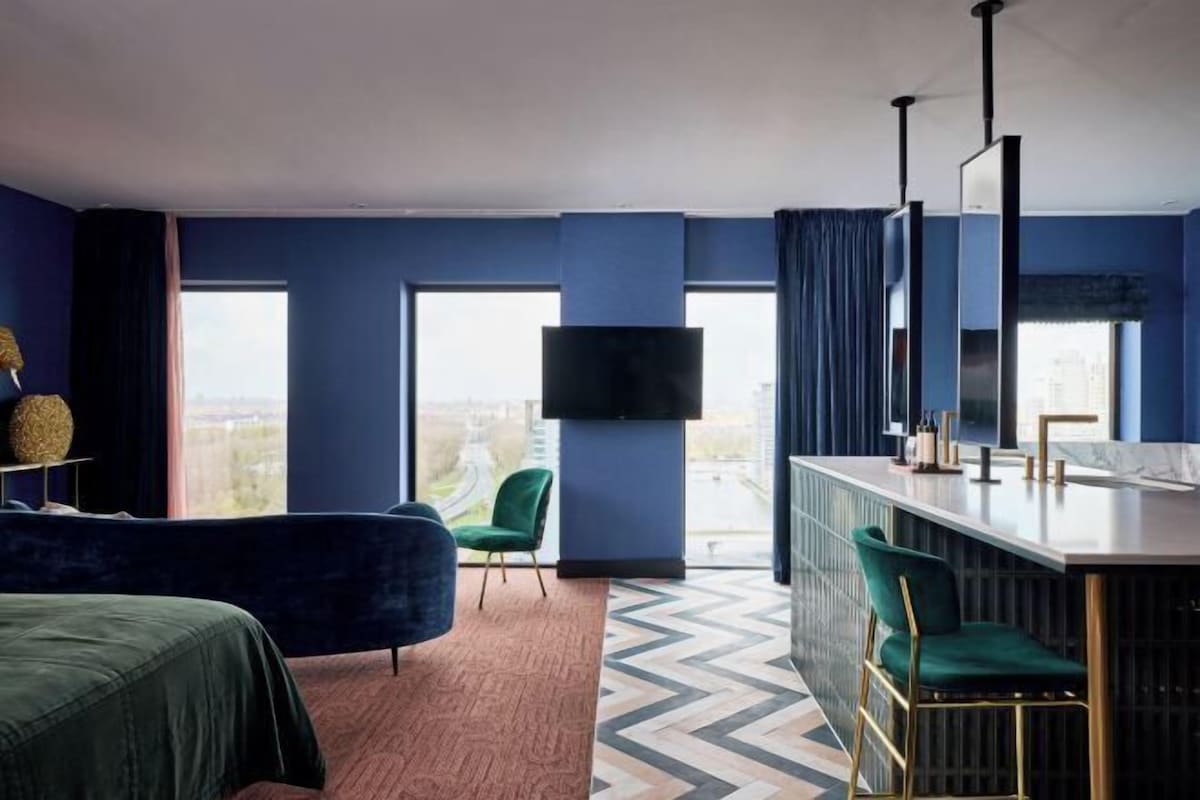 A modern hotel room with a large window, a flat-screen TV, a bar area, and a green chair.