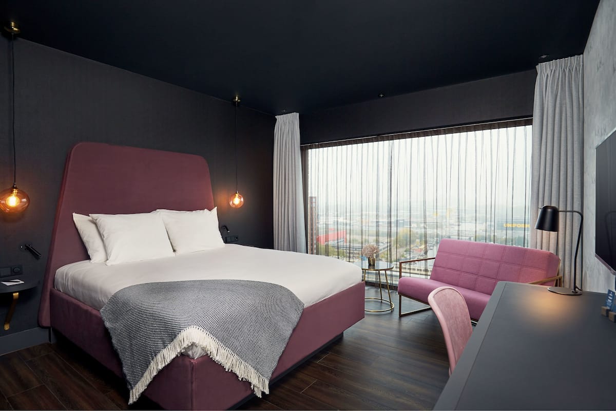 Habitación de hotel moderna con una cama grande, una silla rosa, un escritorio y vistas a la ciudad a través de amplios ventanales.