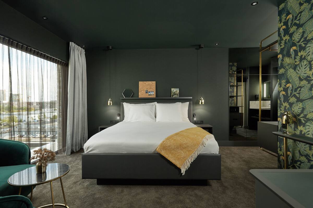 Una habitación de hotel moderna con una cama grande, una mesita, una silla y vistas a la ciudad a través de amplios ventanales.