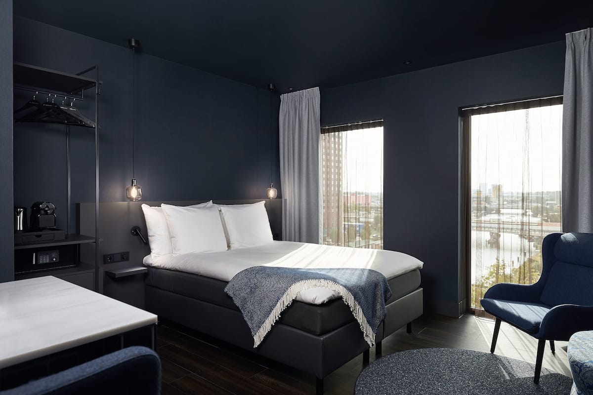Habitación de hotel moderna con una cama grande, un escritorio y una silla. Se ve una ventana con vistas a la ciudad.