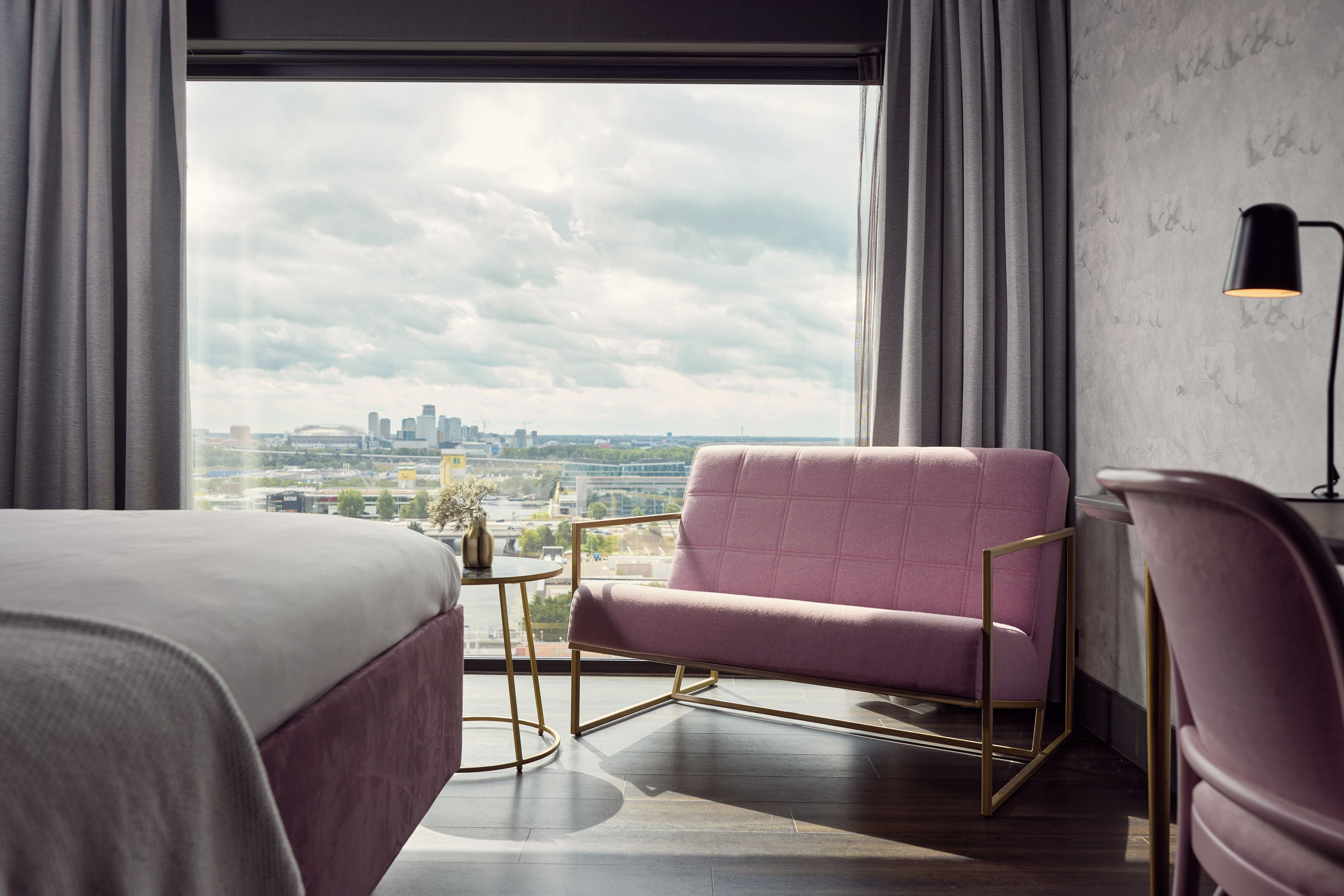 Een hotelkamer met een groot raam, een roze fauteuil, een bed, een klein rond tafeltje en een lamp.