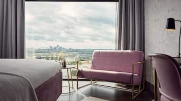 Een hotelkamer met een groot raam, een roze fauteuil, een bed, een klein rond tafeltje en een lamp.