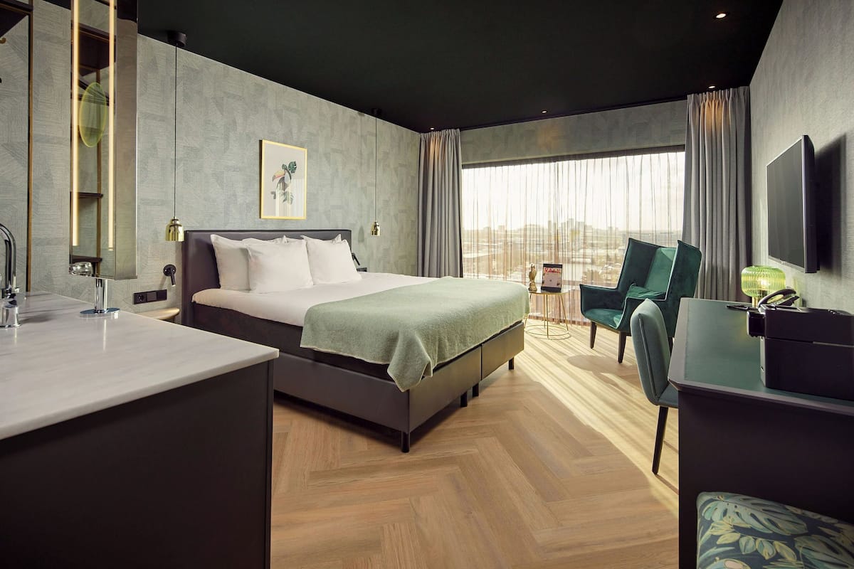 Una habitación de hotel moderna con cama, escritorio, silla, televisor y un ventanal grande.
