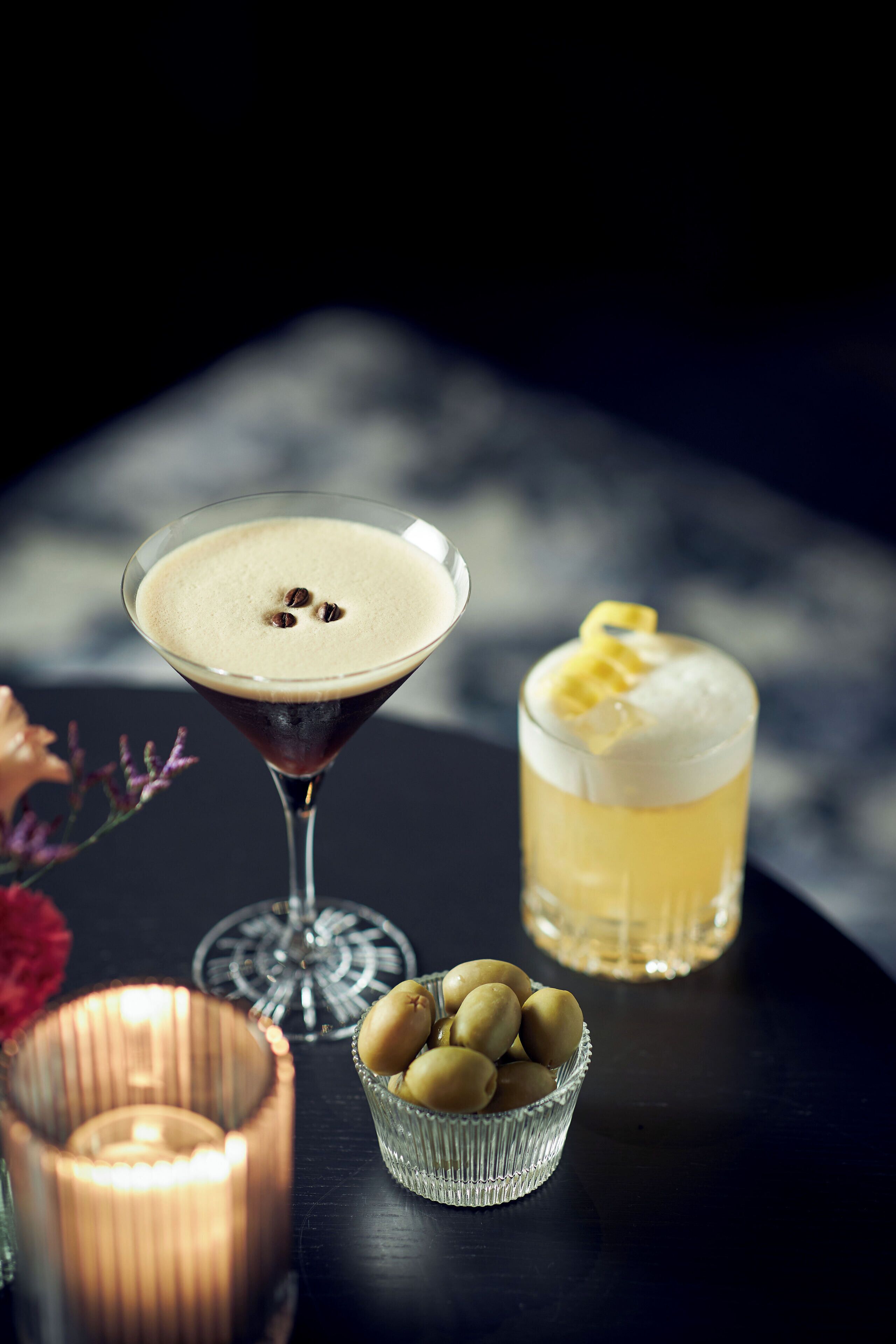A martini glass and a highball glass on a table with a candle and a bowl of olives.