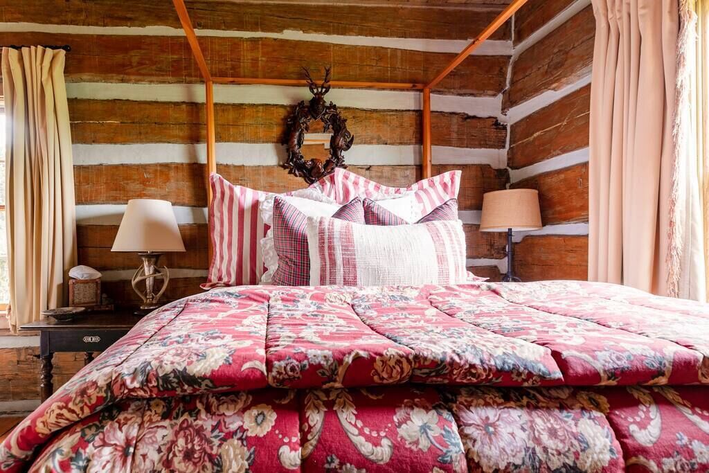 A log cabin bedroom with a four-poster bed, floral bedding, a wooden headboard, a wall-mounted wreath, and a nightstand with a lamp.