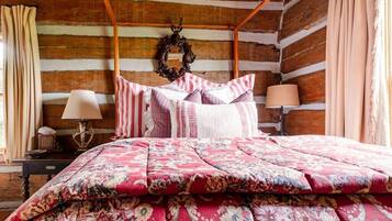 A log cabin bedroom with a four-poster bed, floral bedding, a wooden headboard, a wall-mounted wreath, and a nightstand with a lamp.