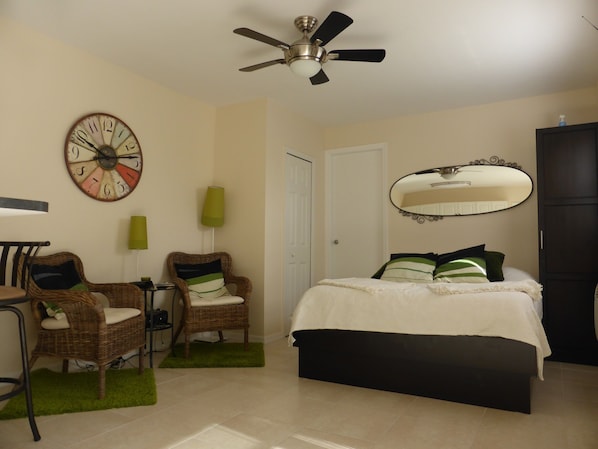 Quarto com uma cama, duas cadeiras de vime, um tapete verde, um relógio de parede, um ventilador de teto, um espelho e uma porta.