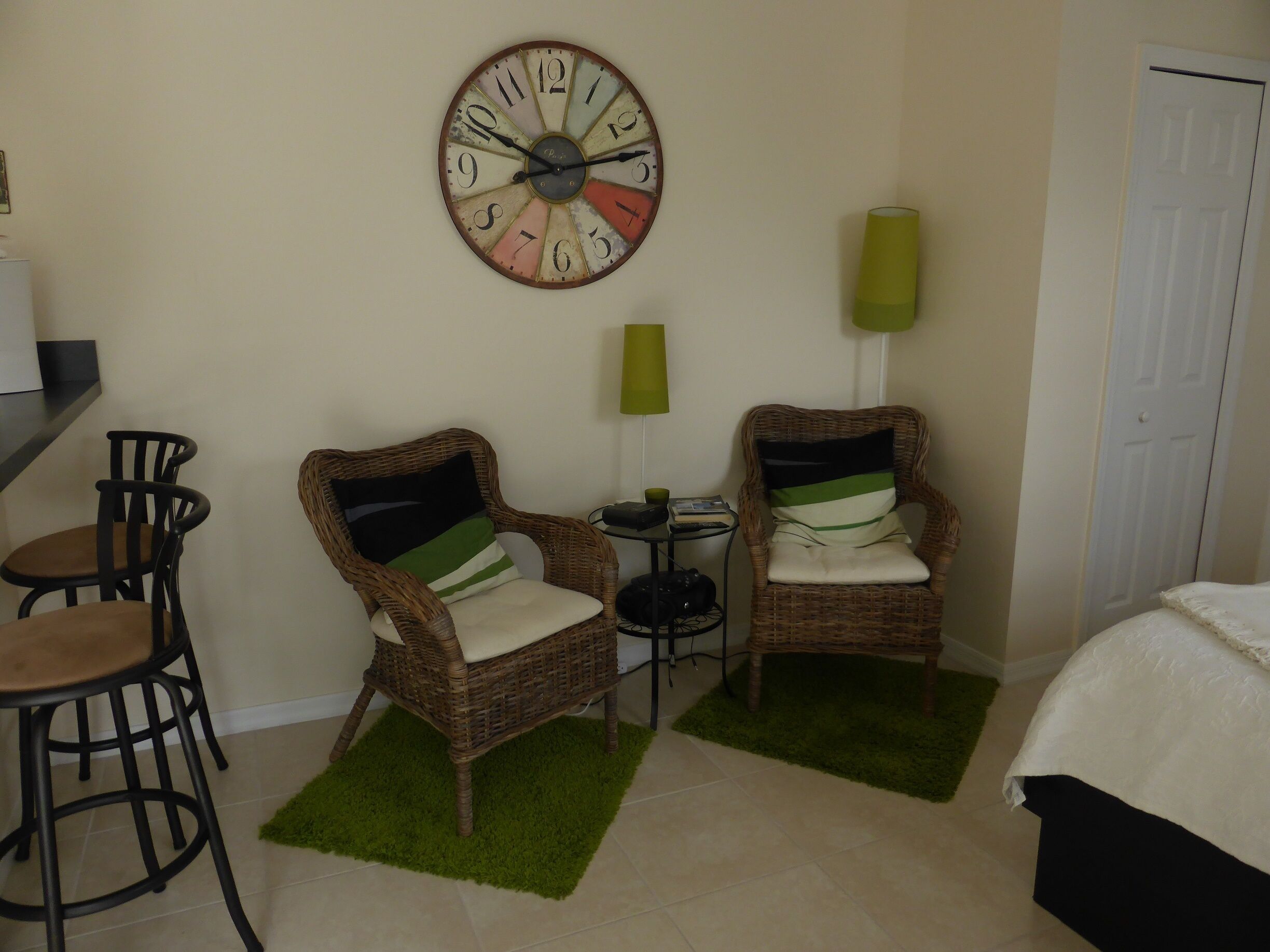 A room with two wicker chairs, a small table, a clock on the wall, and a bed.