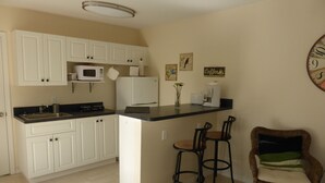 A compact kitchen with white cabinets, a microwave, a refrigerator, and a coffee machine. There are two bar stools and a wicker chair. - Dream holiday at a low price: huge travel discounts now available! (Cape Coral)