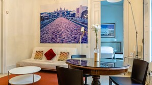 Habitación de hotel con un sofá blanco, una mesa redonda de comedor y un cuadro grande con un paisaje urbano en la pared.