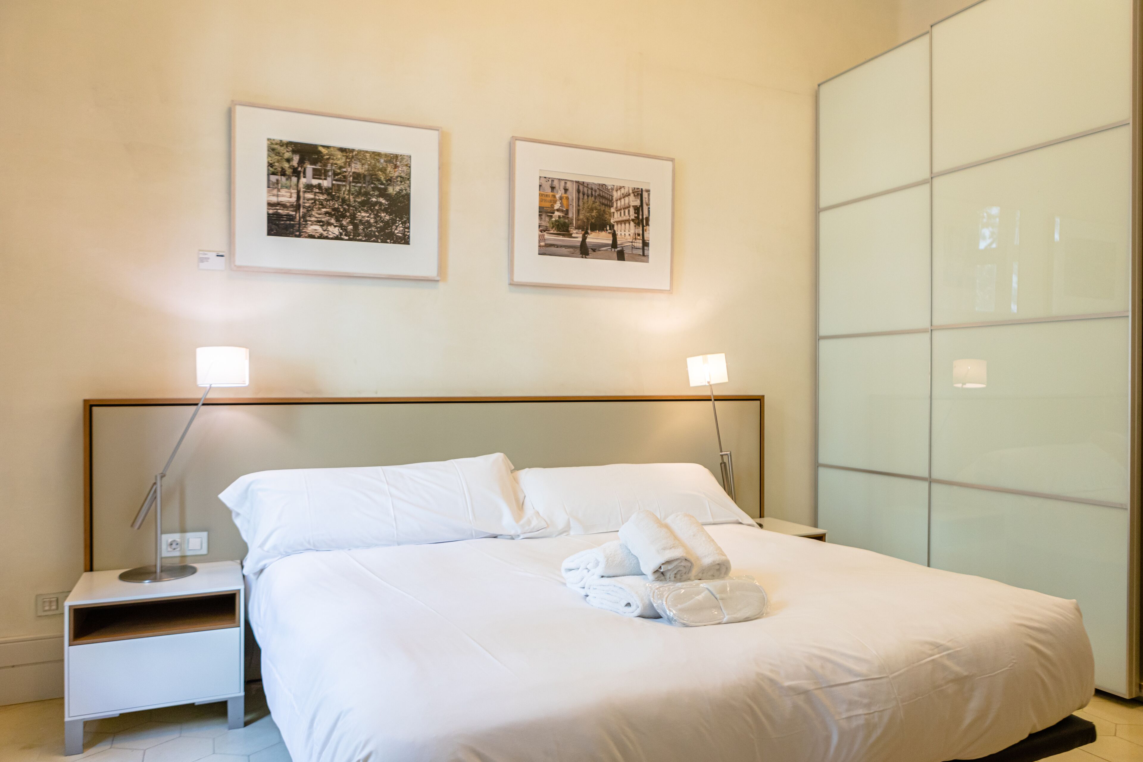 Una habitación de hotel con una cama bien hecha, dos cuadros en la pared, una lámpara de noche y un armario.