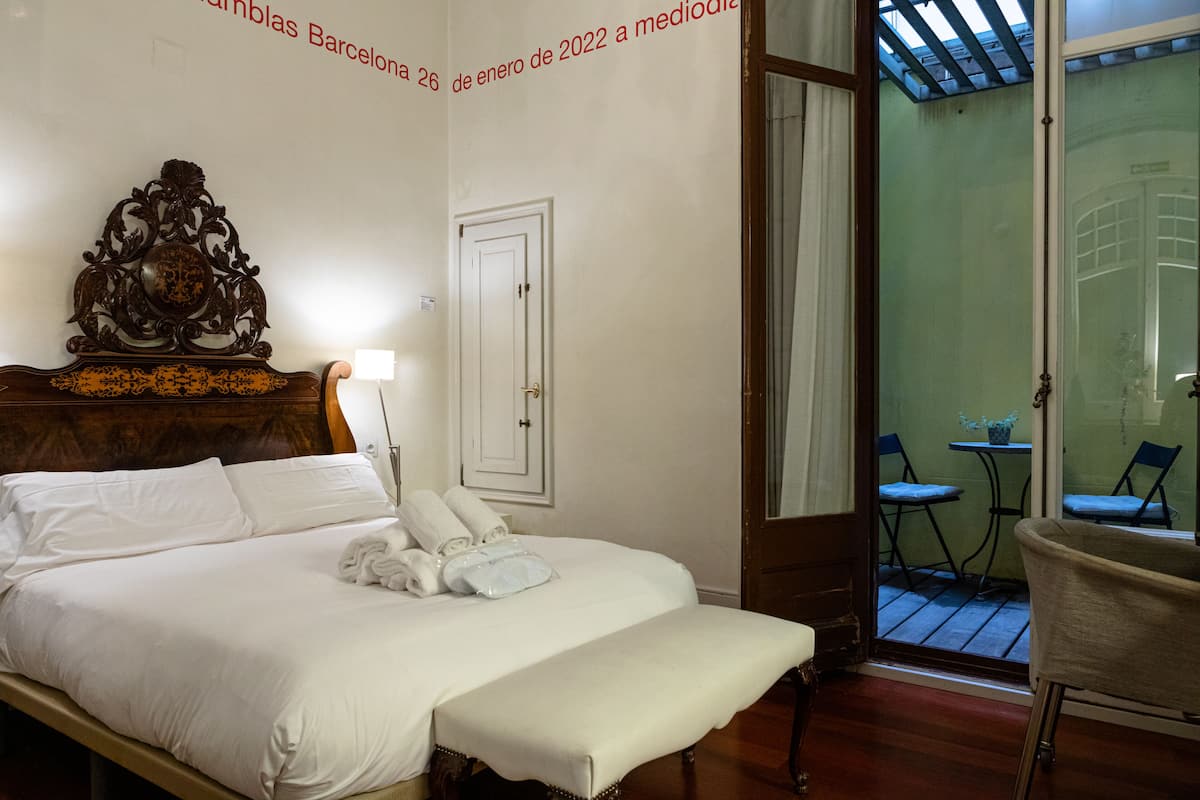 A hotel room with a large bed, a wooden headboard, a bench, a desk, and a view of the outdoor patio area.
