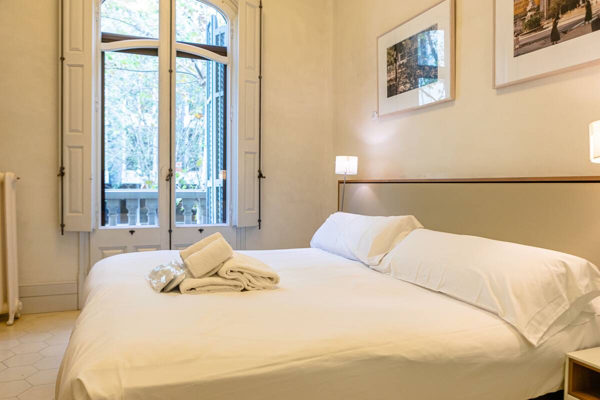 A neatly made bed with white linens and a folded towel, a window with white shutters, and a framed picture on the wall.