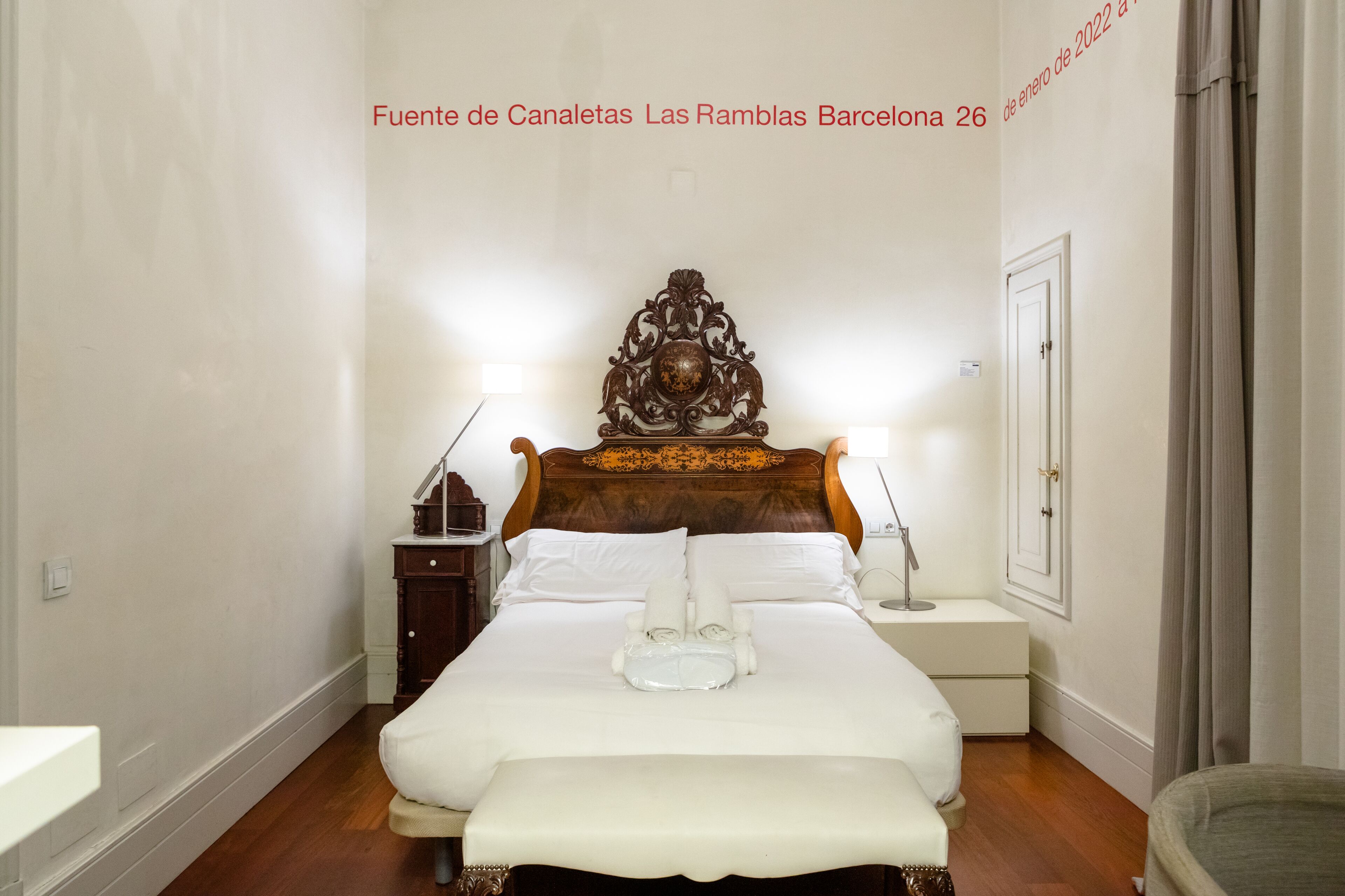 Habitación de hotel con cabecera de madera, ropa de cama blanca, mesita de noche con lámpara y un pequeño mueble auxiliar.