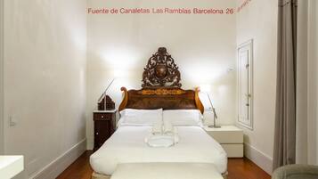 Habitación de hotel con cabecera de madera, ropa de cama blanca, mesita de noche con lámpara y un pequeño mueble auxiliar.