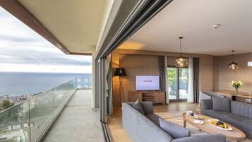 Un salón moderno con un sofá, una mesa de centro y un televisor que muestra la palabra "WELCOME". Un balcón ofrece vistas a un paisaje urbano y al mar.