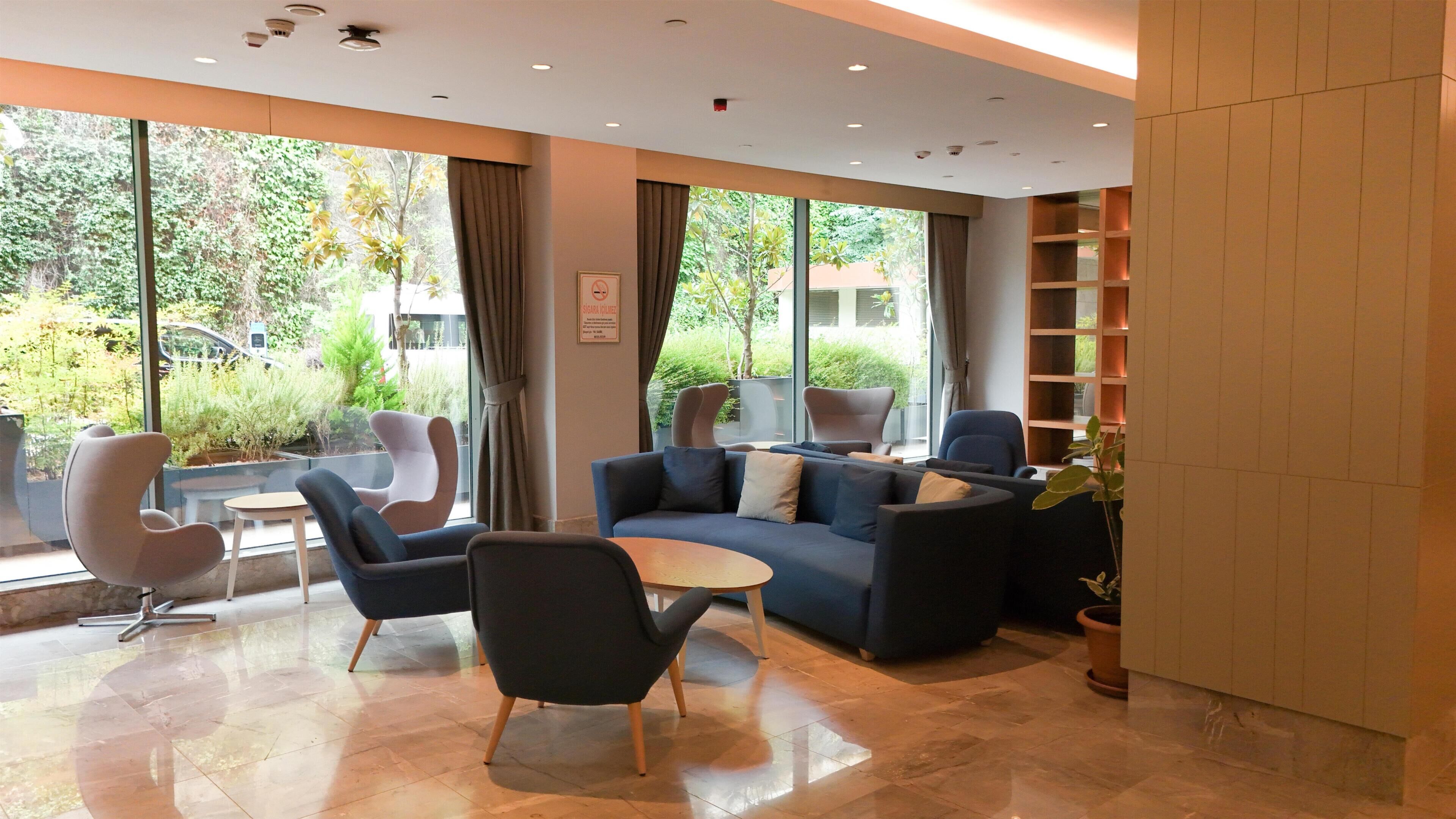 A modern lounge area with a sofa, chairs, and a small table. Large windows provide a view of the outdoors.