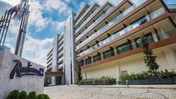 Un hotel moderno de varios pisos con una entrada destacada, un asta de bandera y un patio ajardinado.