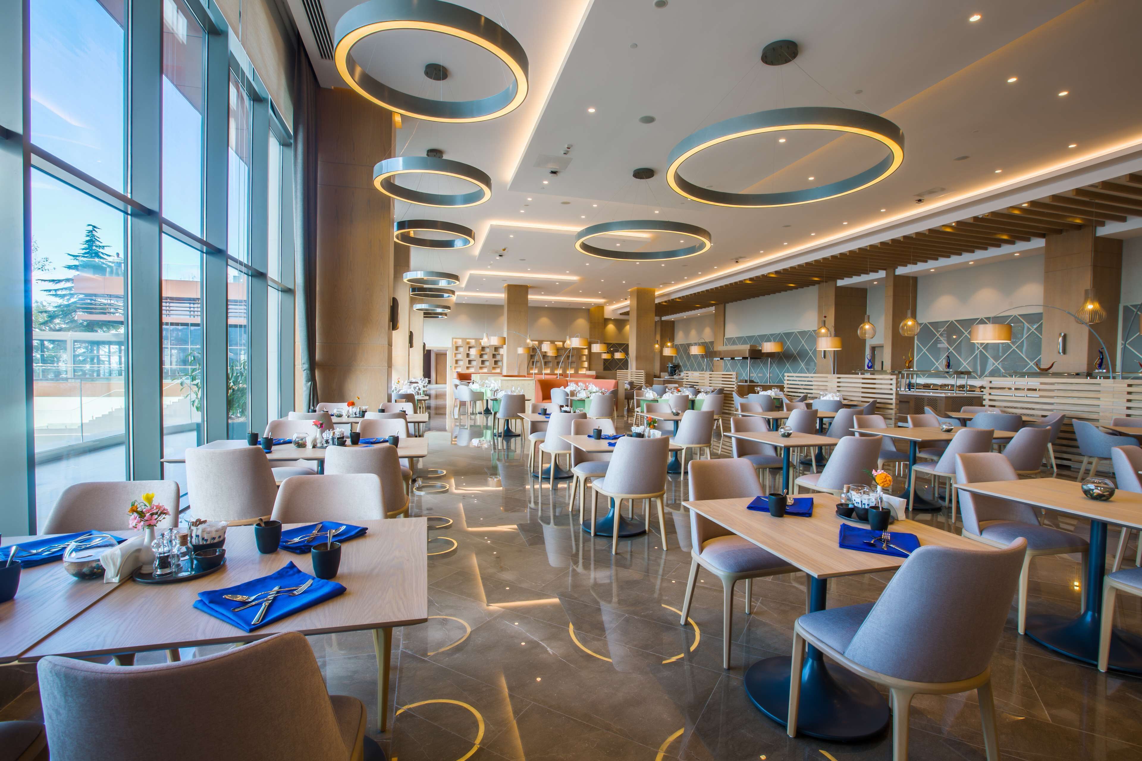 A spacious dining area with multiple tables and chairs, large windows, and modern circular ceiling lights.