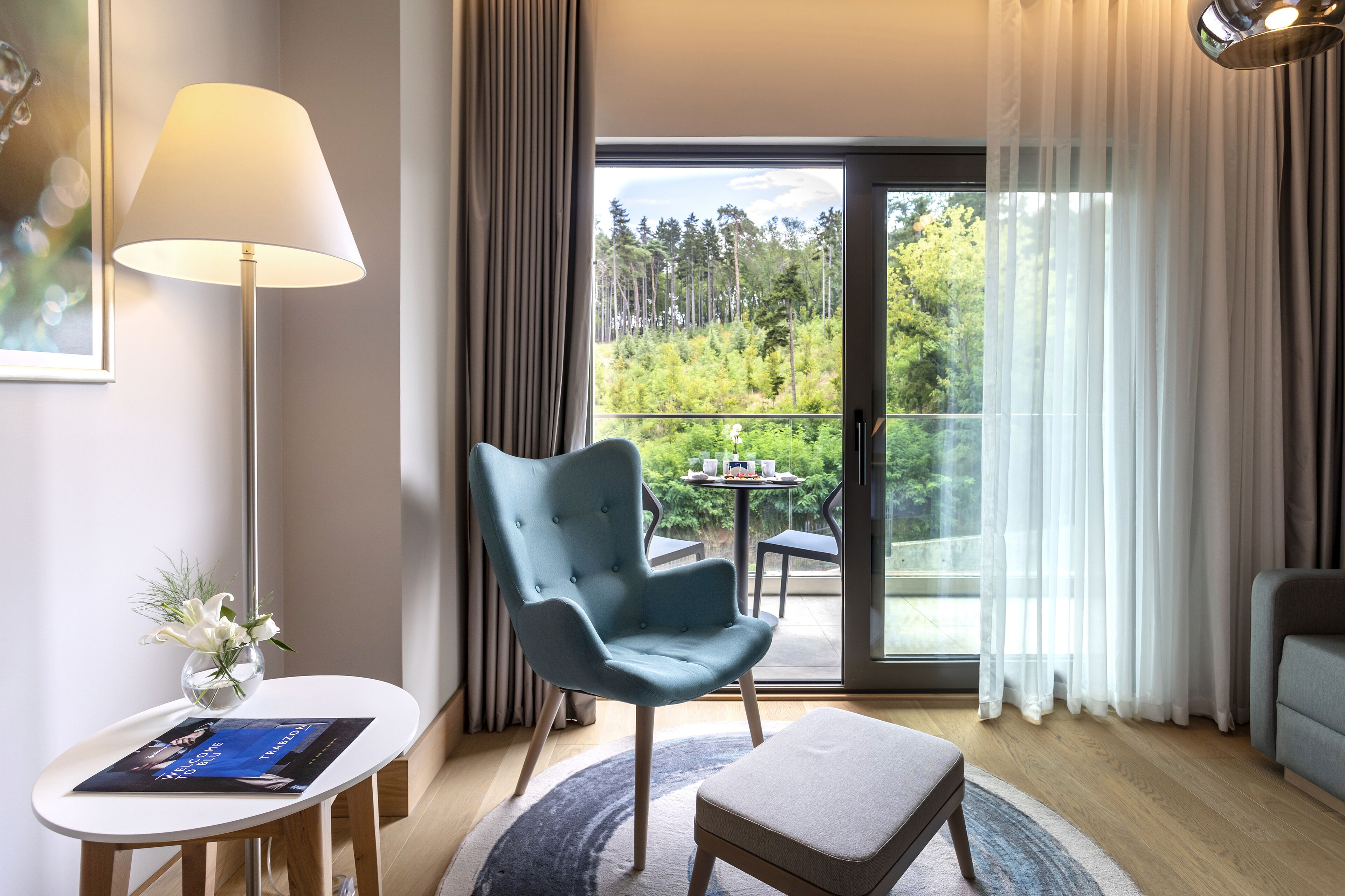 Een moderne hotelkamer met een groot raam, een blauwe fauteuil, een kleine tafel met een bloevas en een lamp.