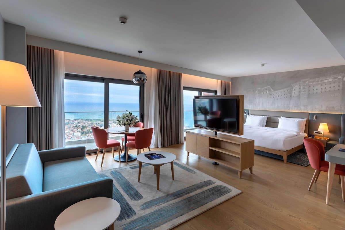 A modern hotel room with a large bed, a TV, a sofa, a dining area, and a view of the city.