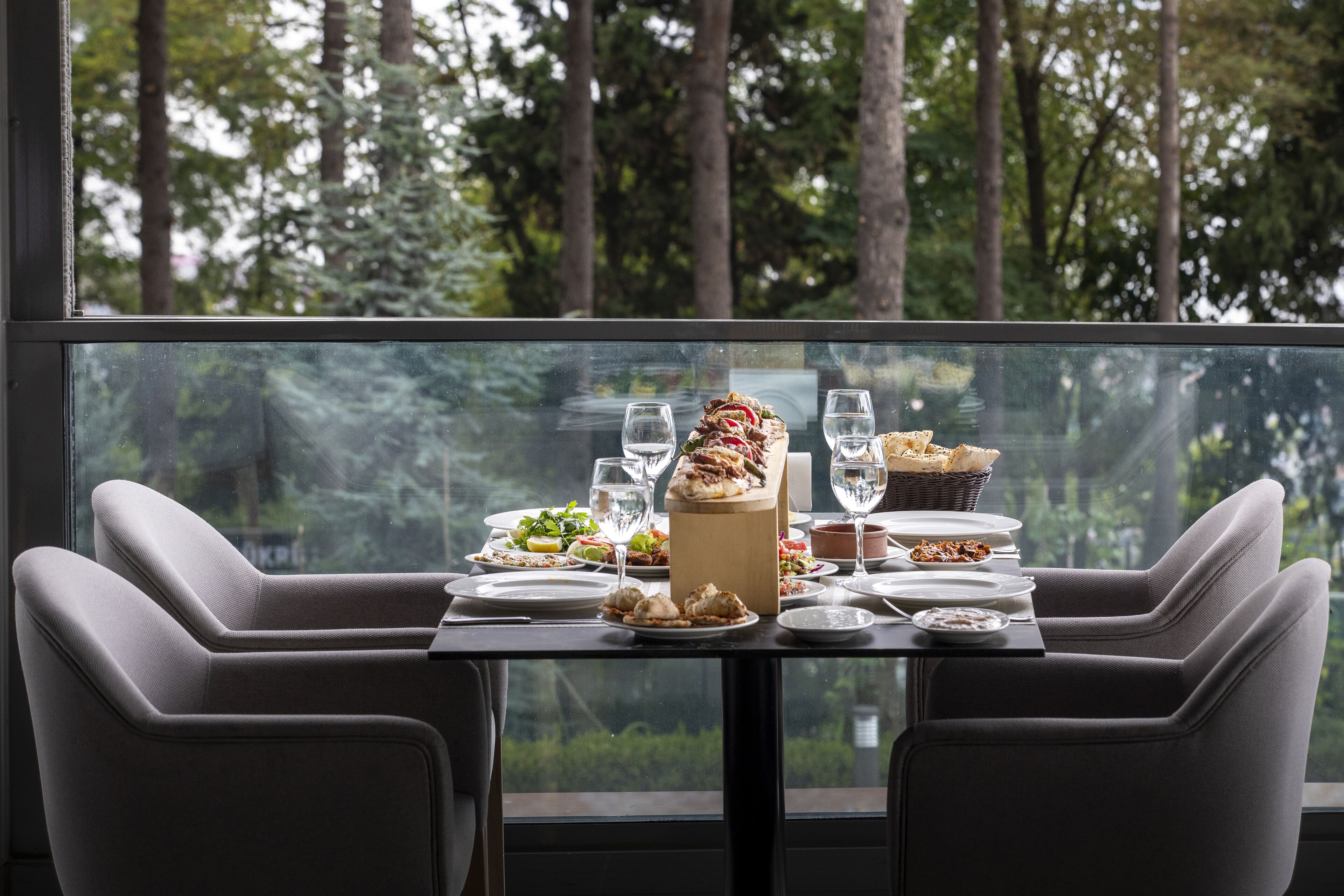 Una mesa de comedor dispuesta con platos, vasos y una tabla de madera con comida, vista a través de un ventanal con un paisaje boscoso de fondo.