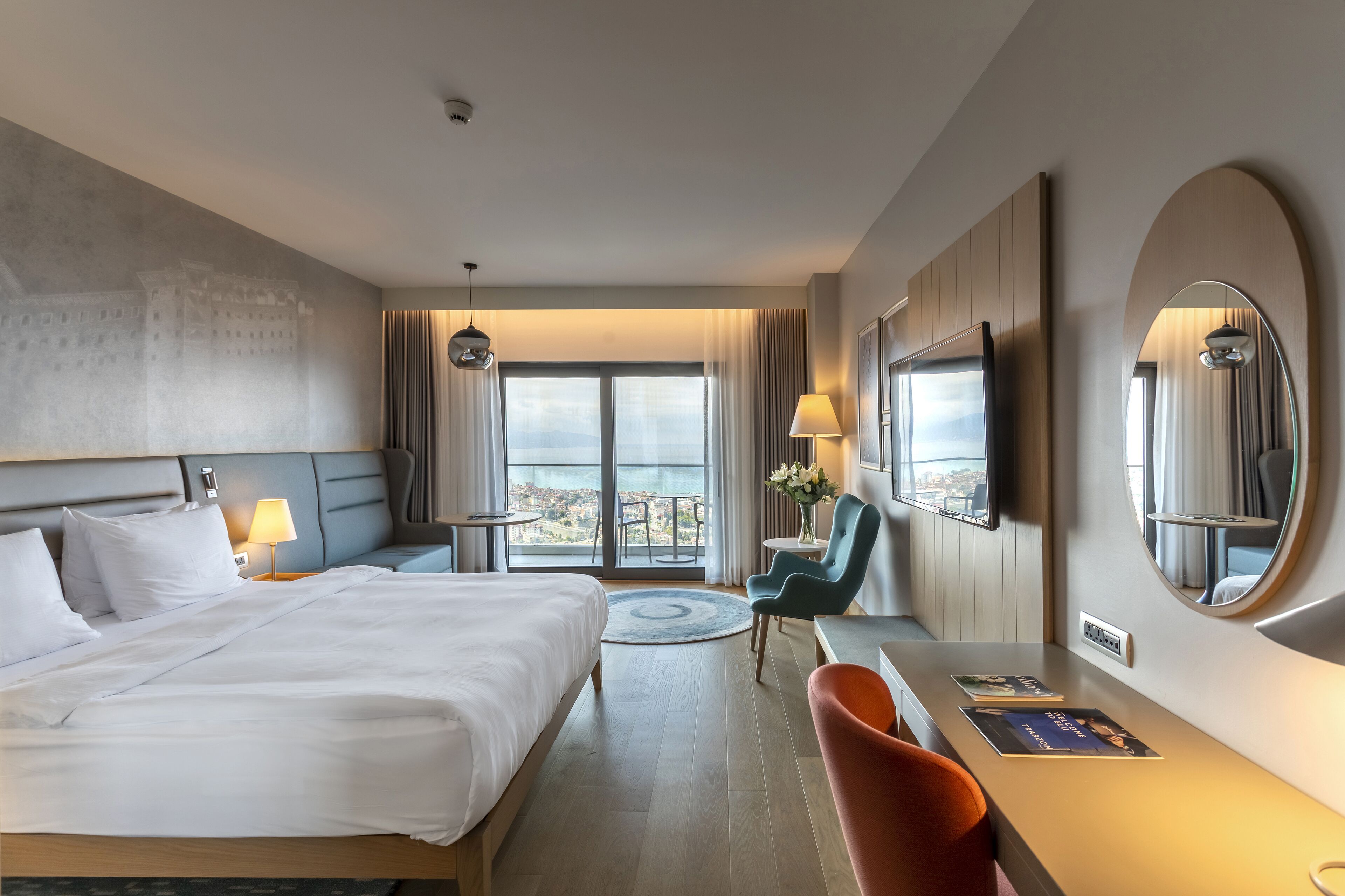 A hotel room with a large bed, a desk with a chair, a round mirror, and a view of the city through the window.