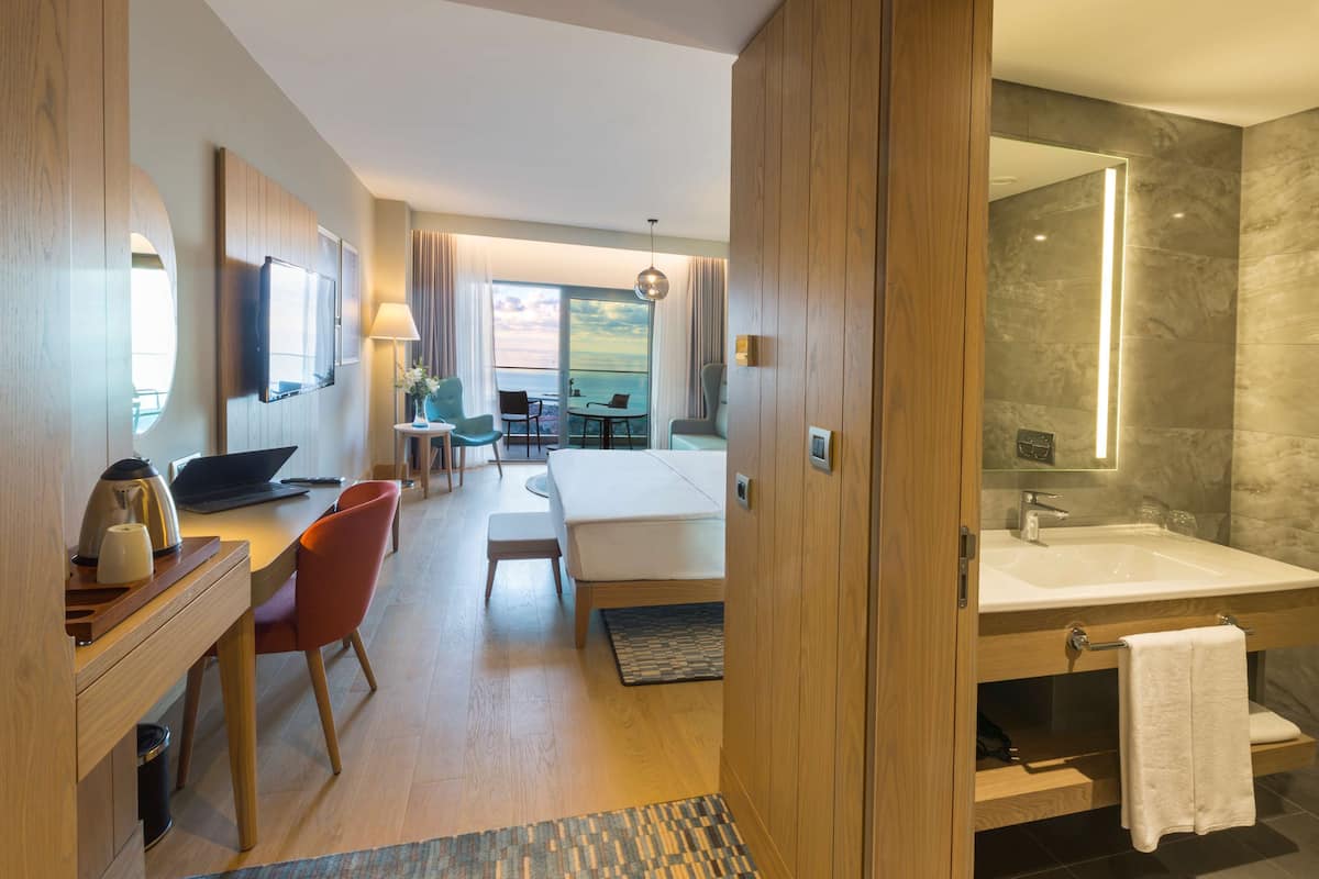 A modern hotel room with a bed, desk, chair, and a bathroom with a large mirror and sink.