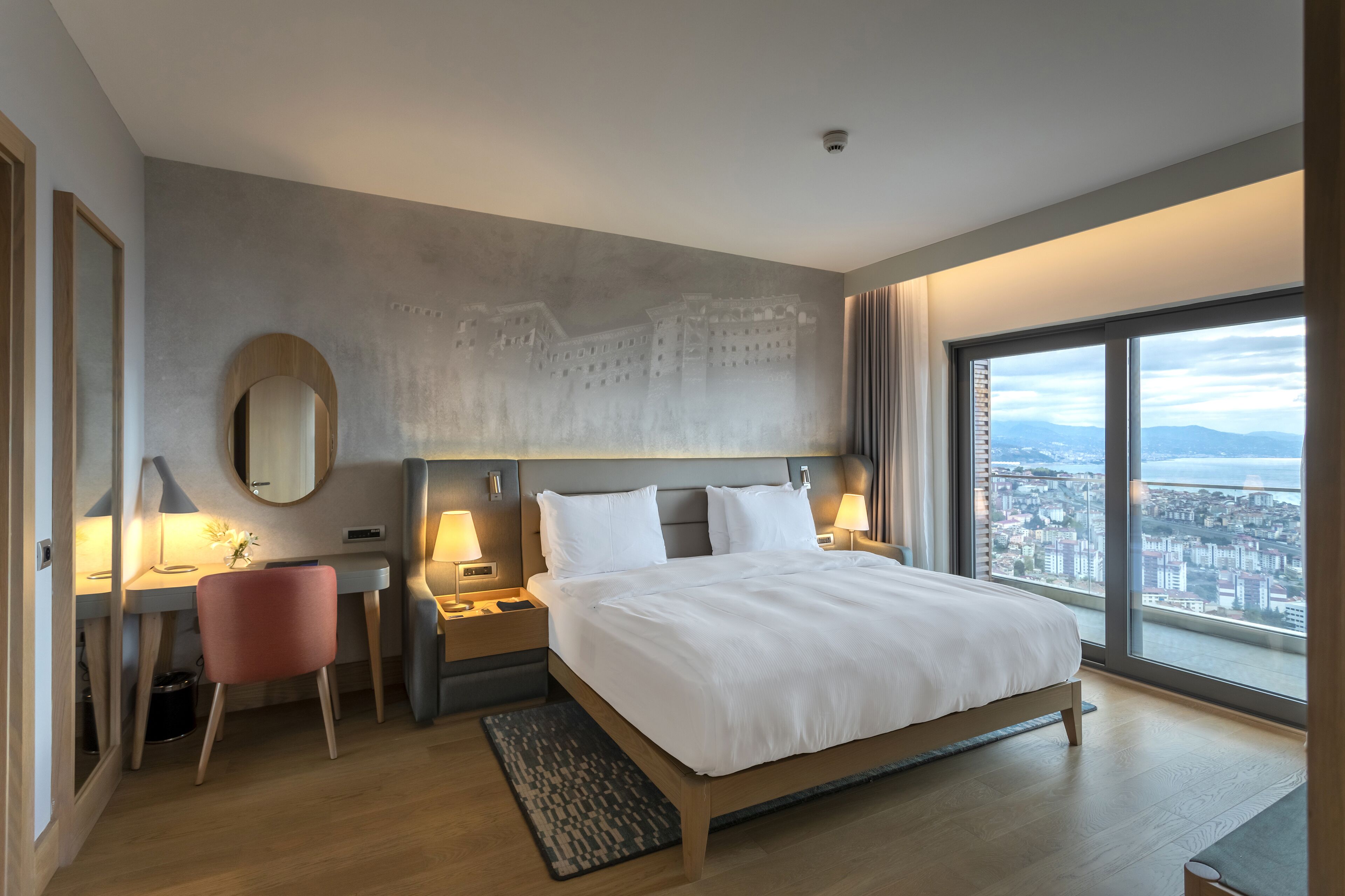 Habitación de hotel moderna con una cama grande, un escritorio, una silla y vistas de la ciudad a través de amplios ventanales.