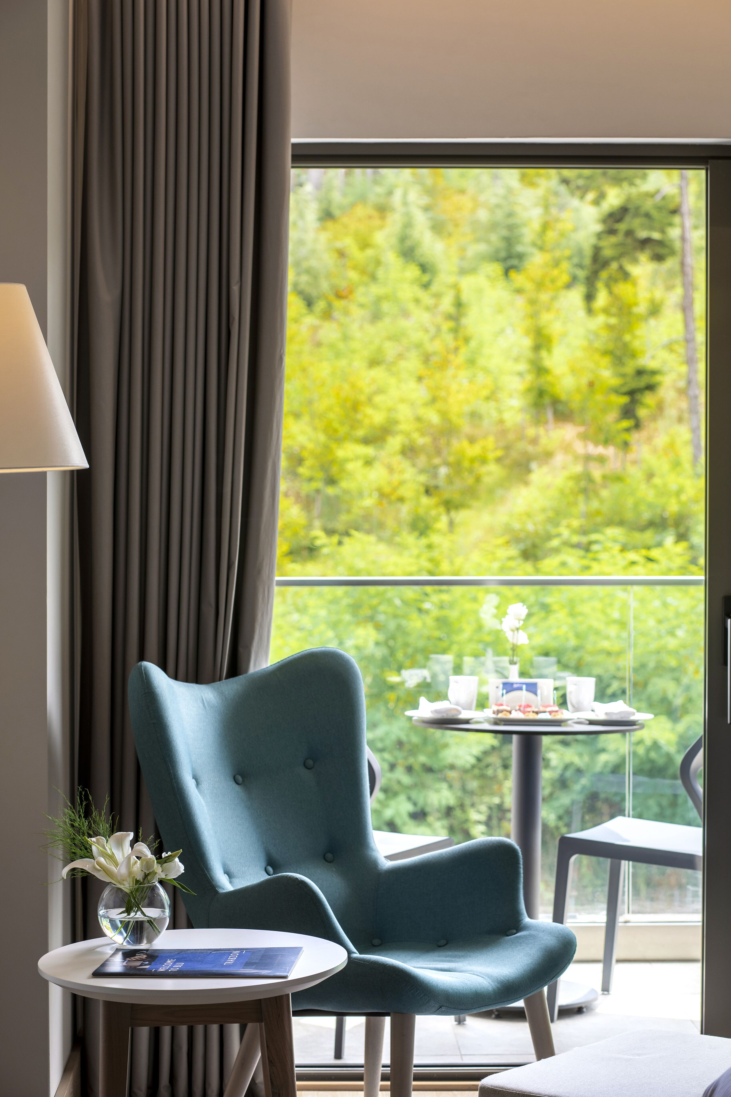 A room with a view of greenery through a window, a blue chair, a small table with a vase of flowers, and a lamp.