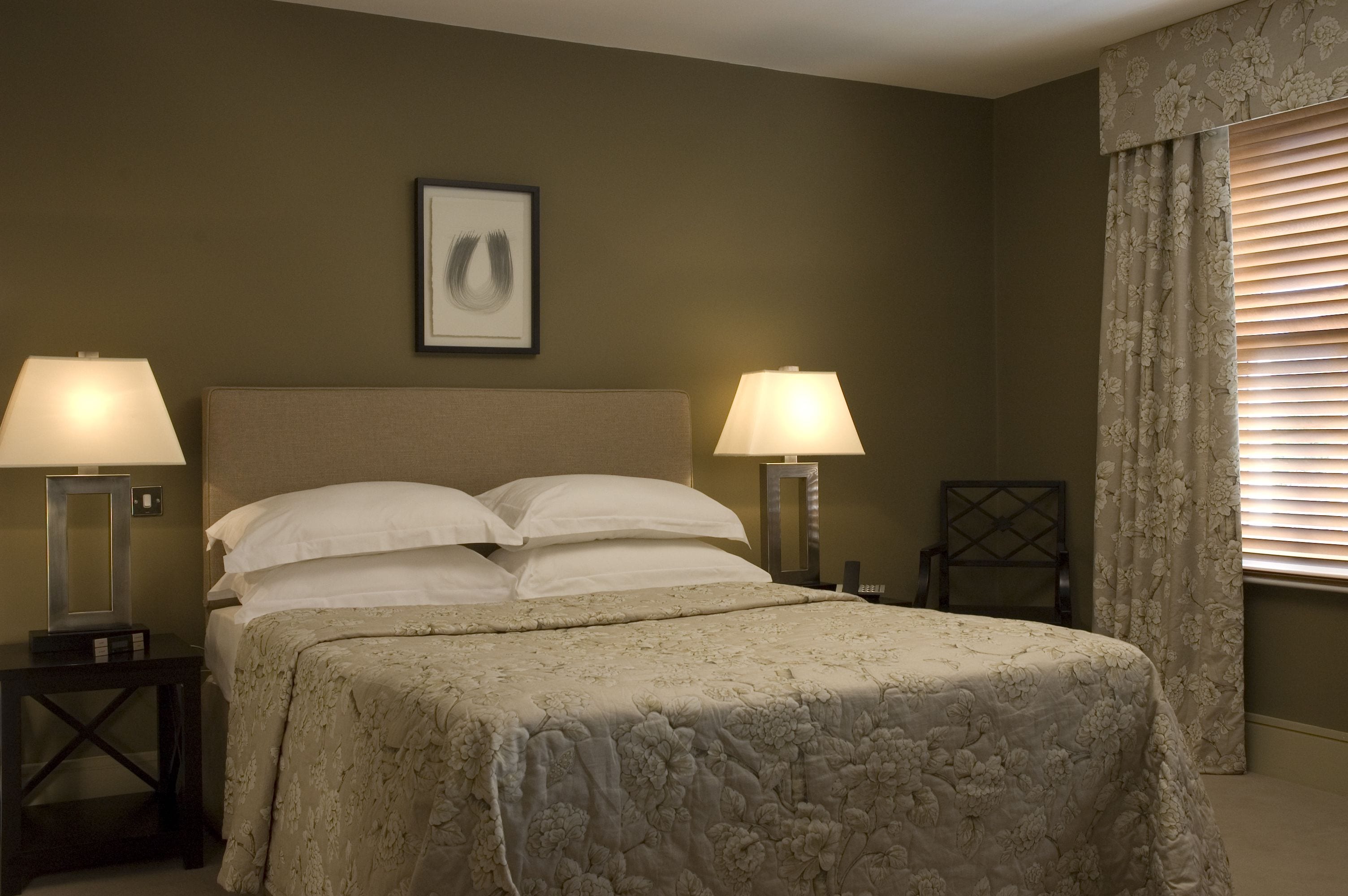 A neatly made bed with a floral bedspread, two bedside lamps, a framed picture on the wall, and a window with floral curtains.