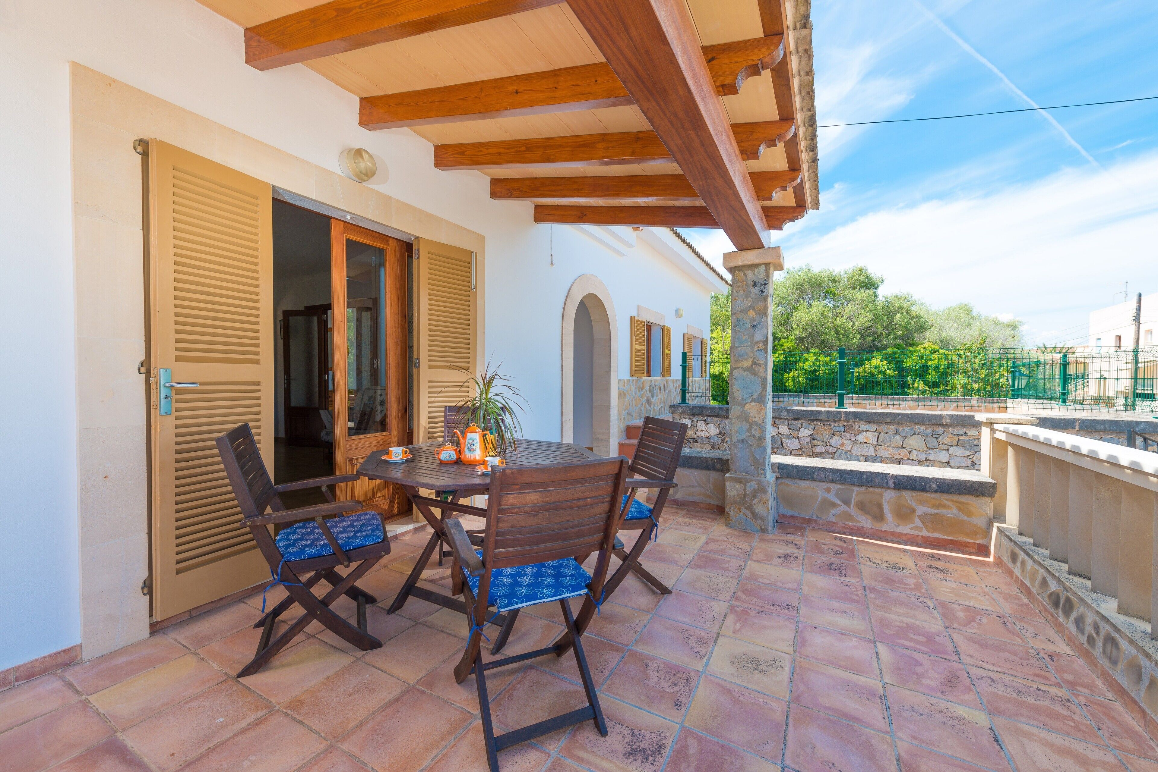 An outdoor patio with a wooden table and chairs, a stone wall, and a view of greenery.