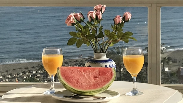 A table with a watermelon, two glasses of orange juice, and a vase with pink roses, set against a view of the ocean. - Tamarindos (Benalmádena)