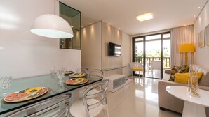 A modern living room with a glass dining table, clear chairs, a sofa, and a TV. - MLW203 Flat at beach Muro Alto 6 people (Ipojuca)