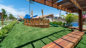 A wooden deck with a slide and play structures, surrounded by green grass and palm trees. - MLW203 Flat at beach Muro Alto 6 people (Ipojuca)