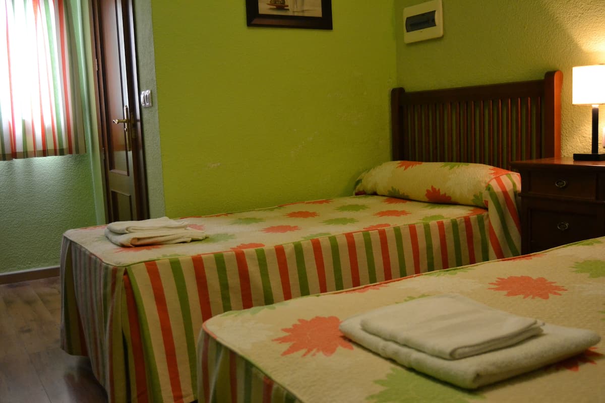 A hotel room with two beds, each with a striped bedspread and a pillow with a floral pattern. There is a nightstand with a lamp and a small table with a folded towel on it.