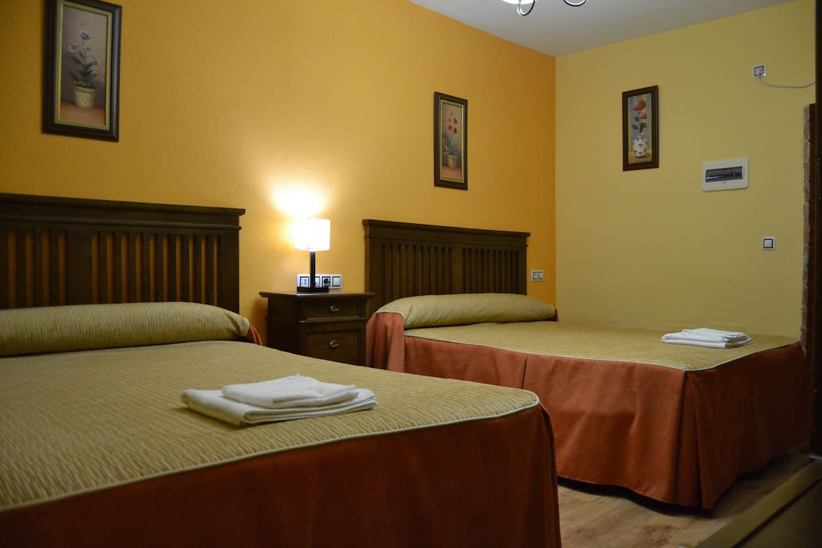 A hotel room with two beds, each with a brown bedspread and a folded white towel on the bed. There is a nightstand with a lamp between the beds. The walls are painted yellow, and there are framed pictures on the wall.