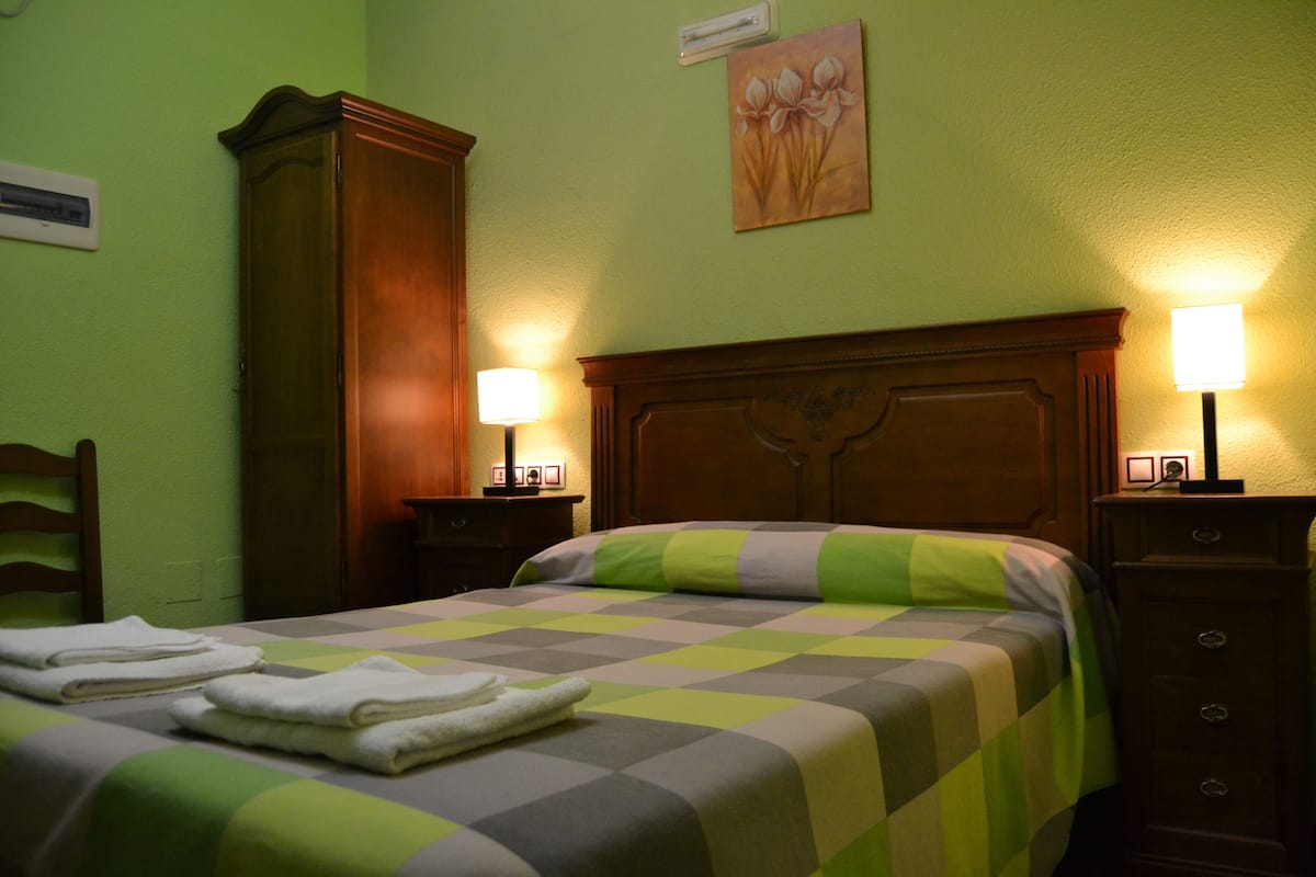 A hotel room with a checkered bedspread, two bedside lamps, a wooden headboard, a nightstand, and a wall-mounted air conditioner.