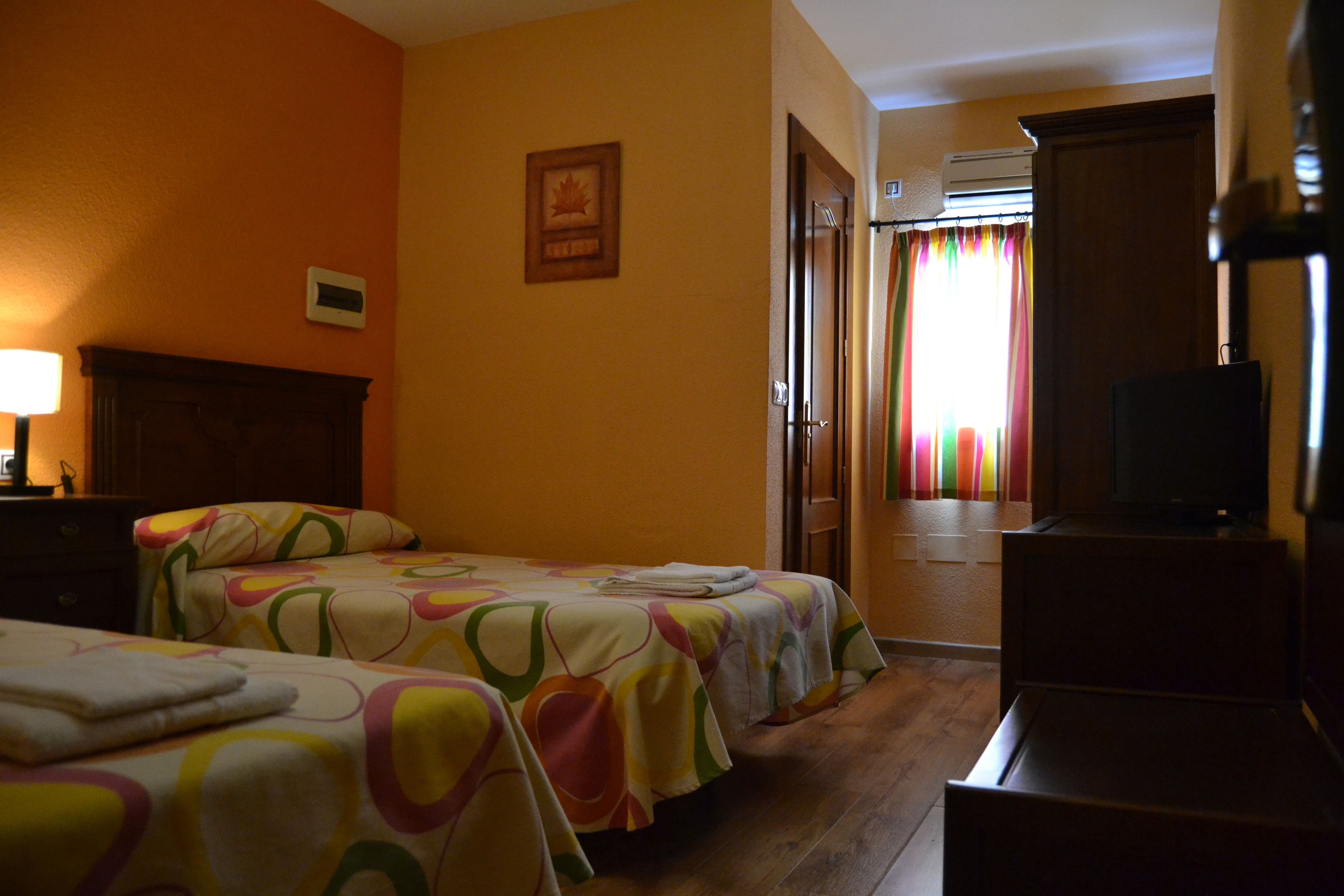 Habitación de hotel con dos camas, cabecero de madera, televisión y puerta que da a un pasillo con cortina colorida.