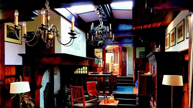 A cozy, wood-paneled room with a fireplace, a chandelier, and a dining area with chairs and a table. - De Zwerver (Naarden)