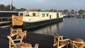A narrowboat docked at a marina with a wooden deck and several wooden chairs. - De Zwerver (Naarden)