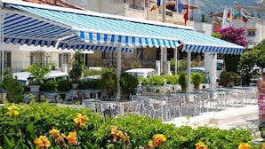 Restaurante com mesas ao ar livre, toldo azul e branco listrado e bandeiras.