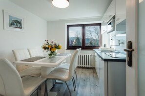 Eine moderne Küche mit weißen Schränken, ein Essbereich mit weißen Stühlen, ein Tisch mit Blumenvase und ein Fenster mit Blick auf Bäume.