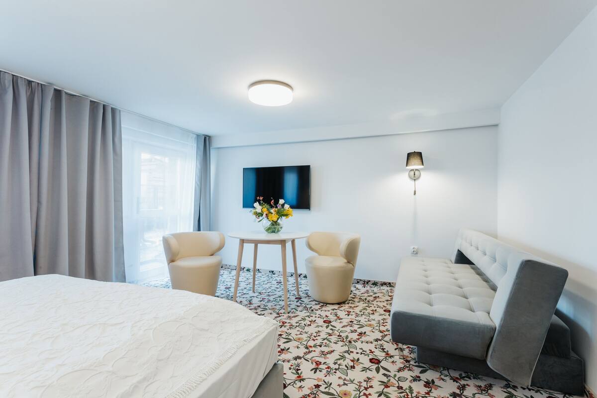 Habitación de hotel con una cama grande, un sofá, dos sillones, una mesita con un florero, un televisor de pantalla plana montado en la pared y una lámpara de pie.