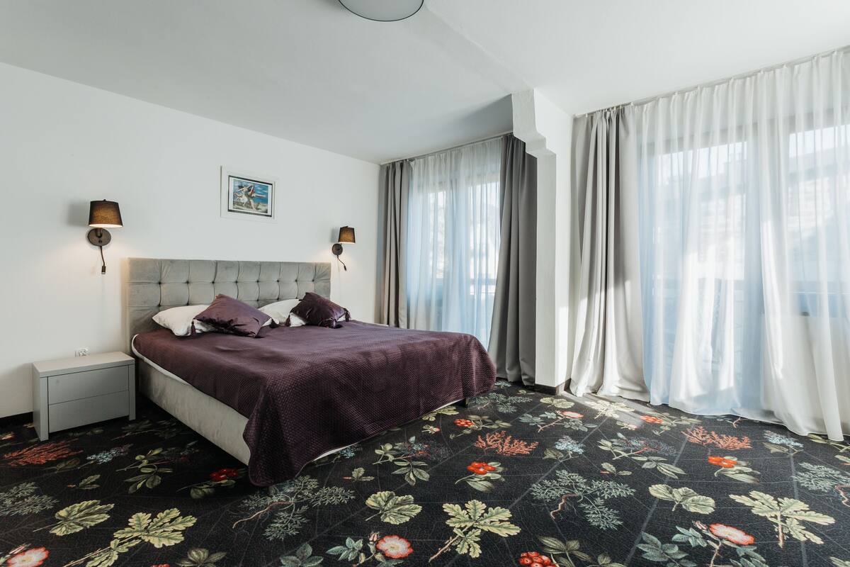 Un dormitorio con cama, mesitas de noche, una lámpara de pared y una alfombra estampada con motivos florales.